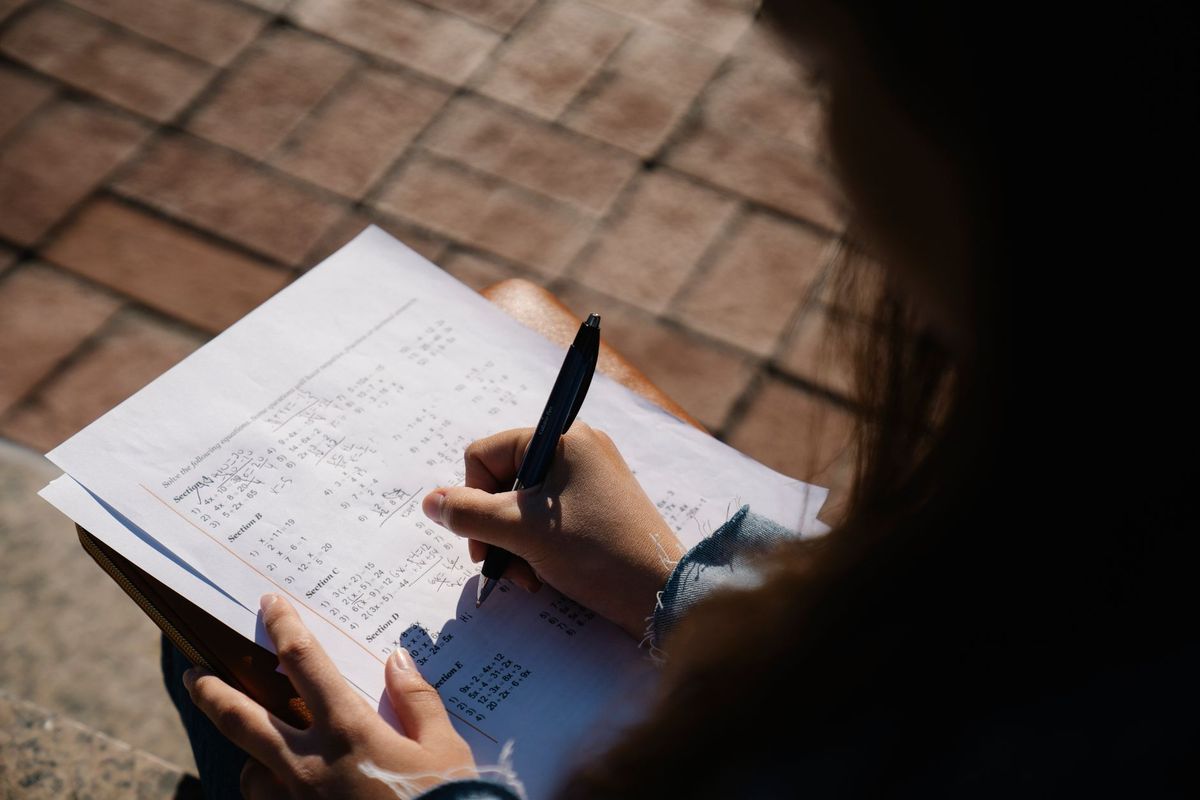 Student holding a pen, reviewing a math worksheet outdoors in natural sunlight